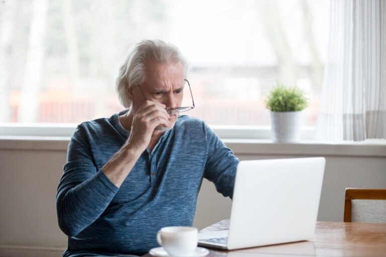 Shocked frustrated senior man taking off glasses looking at laptop